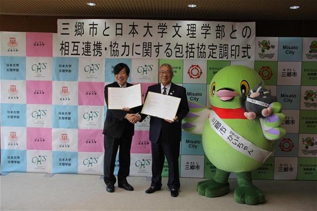 協定書を抱え握手する加藤日本大学文理学部長と木津三郷市長と、市長の右側に三郷市キャラクター「かいちゃん」が立ち、一緒に記念撮影をしている写真