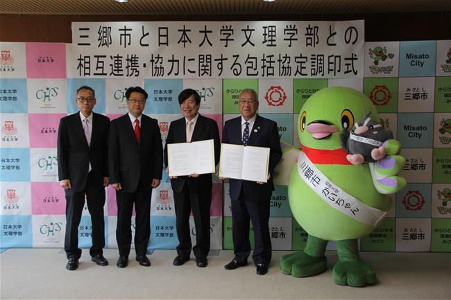 スーツ姿の男性4名と三郷市キャラクター「かいちゃん」が横一列に並び、三郷市長と加藤日本大学文理学部長が協定書を持って記念撮影をしている写真