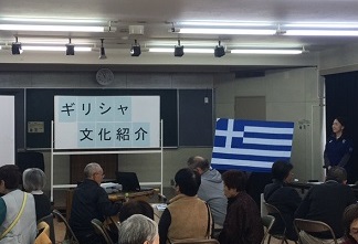 ギリシャ国旗が設置されている公民館内で、ホワイトボードに「ギリシャ文化紹介」の紙が貼られ、前方で一人の女性が話をしている様子と、椅子に座っている参加者たちが話を聞いている写真