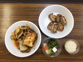 白いお皿やプラスチックの透明カップに、パン料理や煮込み料理、ニンジンや葉野菜を使ったサラダがテーブルに並べられている写真