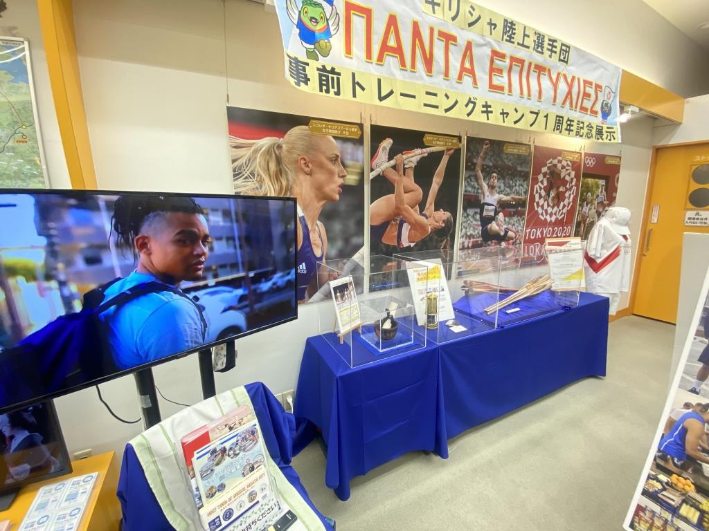 壁に選手が写った大きなポスターが5枚貼られ、映像が流れているテレビの右側に聖火リレートーチやユニフォームなどが展示されている写真
