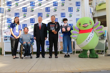 三郷市のバックボードの前で木津市長や、かいちゃん つぶちゃんと一緒に並んで記念撮影をする4人のゲストの写真