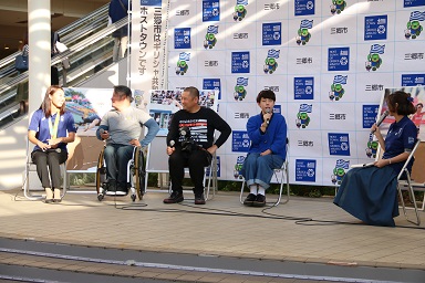 三郷市のバックボードが設置された屋外の会場で、座って話をしている4人のゲストのかたと、女性司会者の写真