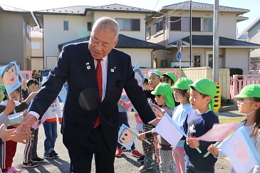 手作りの旗を手にした園児たちが両側から列を作り、中央を歩く木津市長と握手を交わしながら出迎えている写真