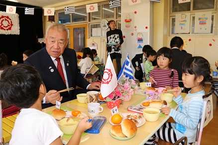 ギリシャと三郷市の旗が飾られたテーブルで会話をしながら給食を食べている木津市長と園児の写真