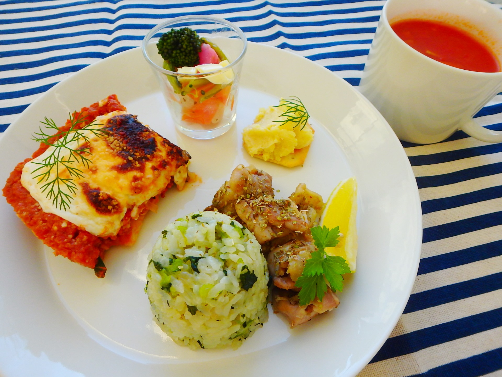 ストライプのテーブルクロスの上に、ワンプレートに綺麗に盛付けされたギリシャ料理（じゃがいもとなすの三郷風ムサカ、小松菜ピラフ ギリシャ風 on 鶏肉のスブラキ、フェタチーズの旬野菜サラダ、タラモサラタ、レンズ豆のミネストローネ）の写真