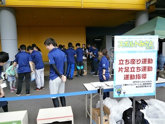 イベント会場外に「立ち座り運動 片足立ち運動 運動指導」と書かれたボードが設置され、青いTシャツを着用したスタッフが集まっている写真