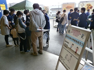 屋外の会場に「自分の体力年齢を知ろう」と書かれた看板が設置され、スタッフや来場者で賑わっている写真