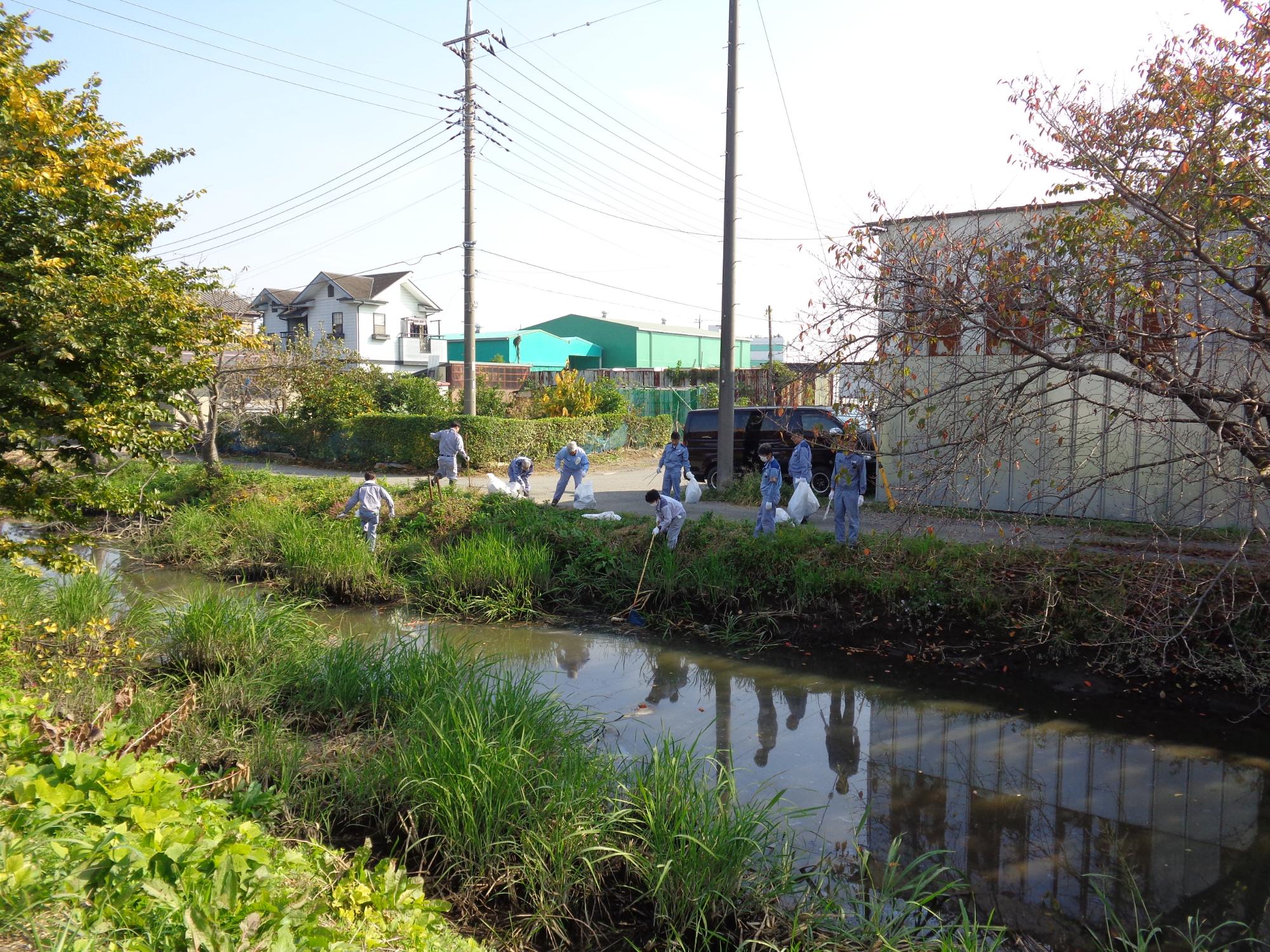 袋を持った男女数名が河川敷のゴミを火ばさみや網を使って拾っている写真