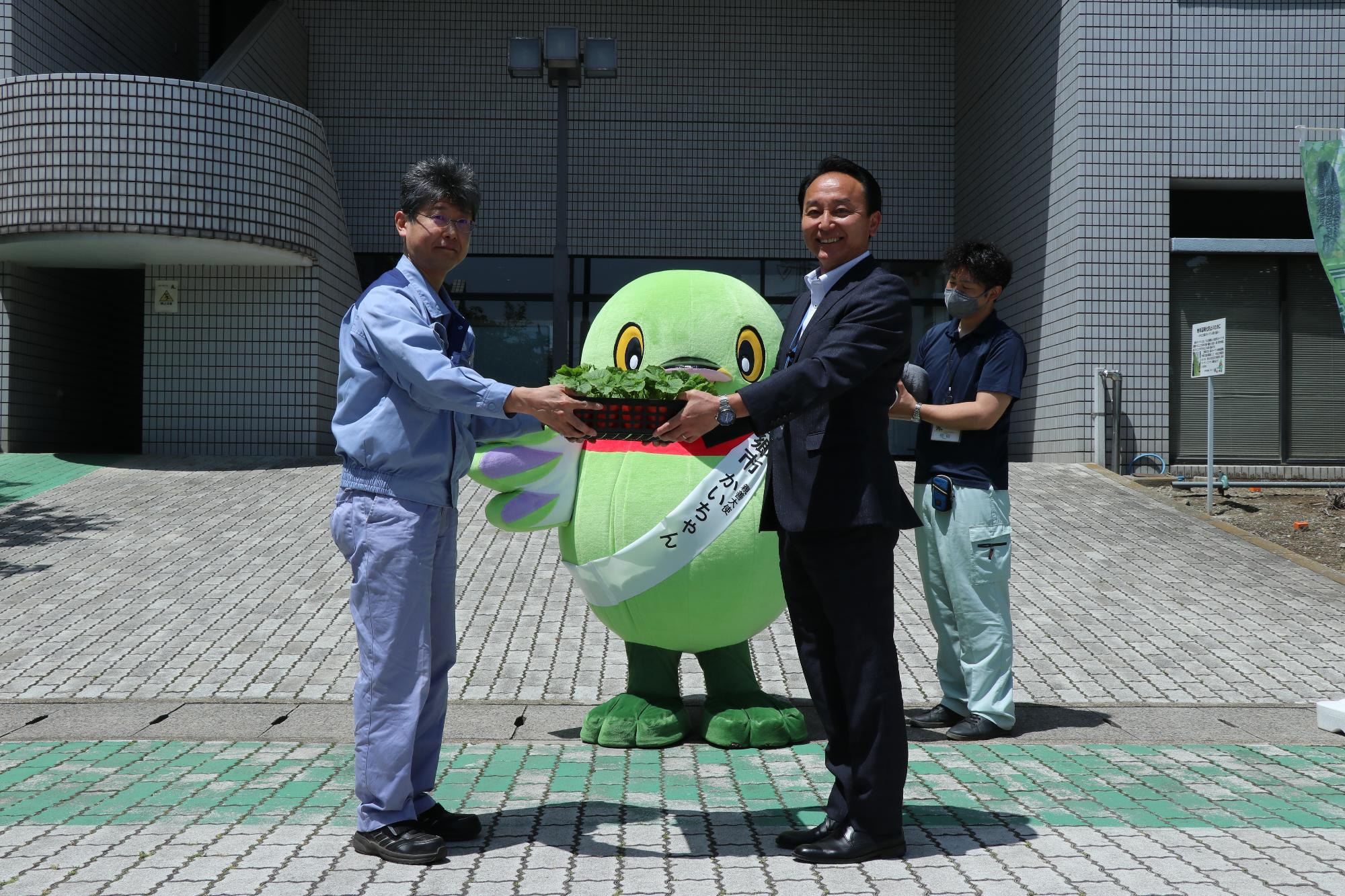 三郷市キャラクターかいちゃんが見守る中、スーツ姿の2名の男性が向かい合い、ゴーヤ苗を一緒に持っている写真