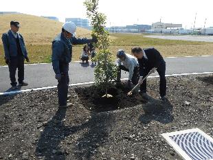 3名の男性が道路脇の敷地に記念樹を植えており、その様子を道路側から見ている帽子を被った男性や、子ども連れの女性を写した写真