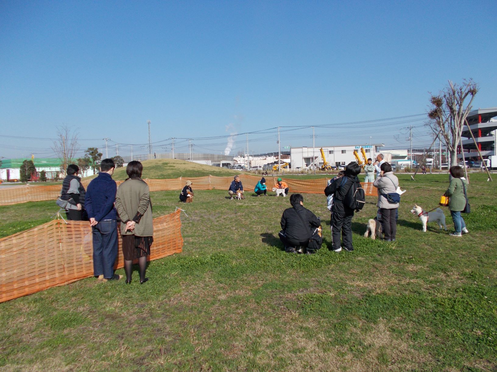 芝生の上に設置されたオレンジ色の囲いの中に犬をつれた人達が円になり講師の話を聞いている様子を正面から撮影した写真