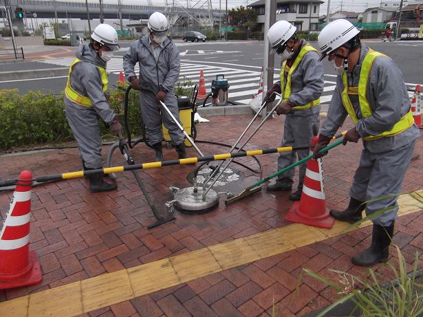 ヘルメットと作業着を着用した4人の作業員が、交差点近くの歩道で高圧洗浄による除染作業をしている写真