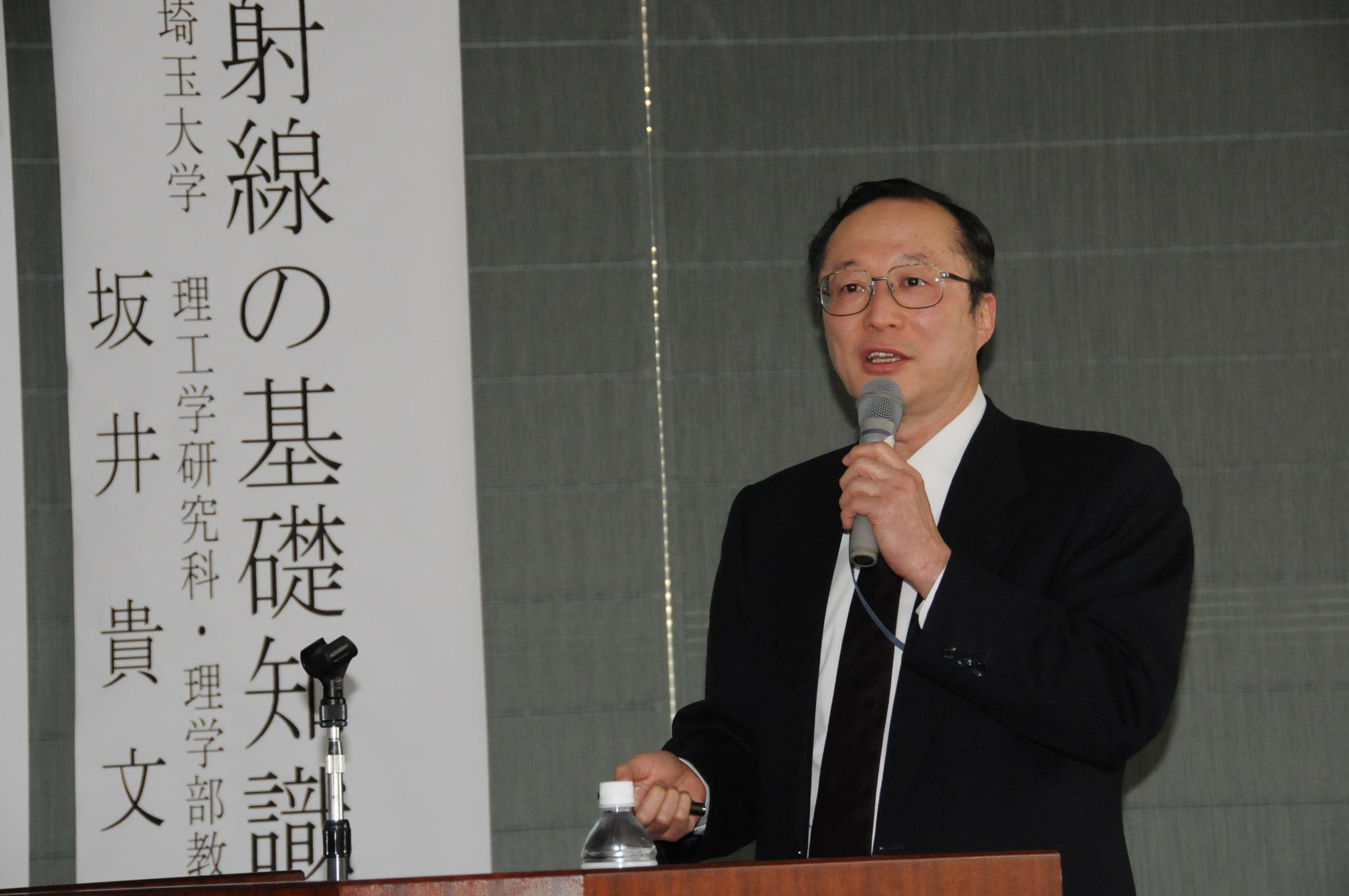 登壇に立ってマイクを持ち、放射線の基礎知識について講演する坂井氏の写真