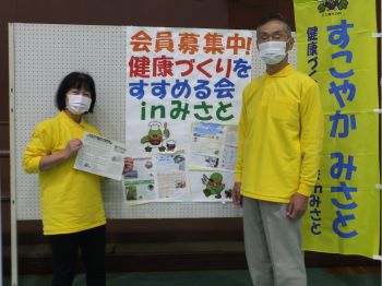 「会員募集中！健康づくりをすすめる会inみさと」と書かれたポスターの前に立つ、黄色い服を着た男女2人の写真