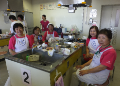 調理台の周りの椅子に座ってカメラ目線で微笑んでいる5人の女性スタッフの写真