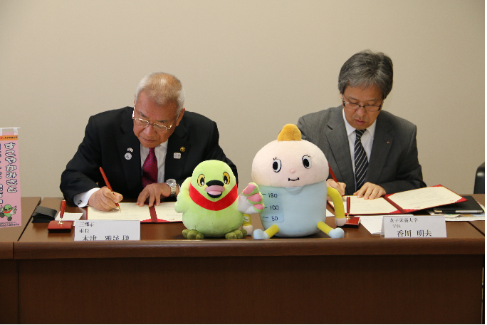 三郷市長と香川女子栄養大学学長が並んで机に座っており、協定書にサインをしている様子の写真