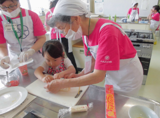 袖口に「ヘルスメイト」と書かれたピンクのTシャツ姿の女性が、参加者の女の子と一緒に調理作業をしている写真