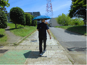 日差しの強い中、男性が小さめの青い日傘をさし、リュックを背負って歩いている姿を背後から撮った写真