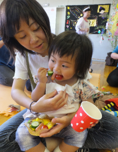 女性の膝の上に座った子どもが串に刺さっているトマトやウィンナー、きゅうりを口へ運んでいる写真