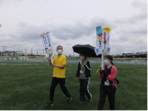 中央に日傘をさした女性、両側に熱中症予防啓発の立て札を持った男女が並んで会場内を歩いている様子の写真