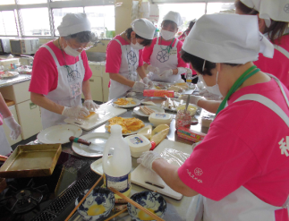 ピンクのTシャツに白の三角巾とエプロンを身に着けた女性たちがまな板の上に食材を置き作業をしている写真