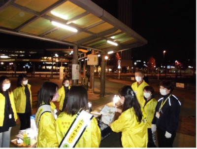 街頭キャンペーンに参加した黄色い上着を着た10名ほどの参加者が駅前のロータリー付近に集まり打ち合わせを行っている様子の写真