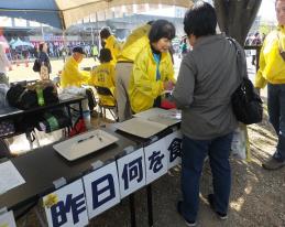 テントの中に黄色い上着を着た会員数名が作業をしており、その内の1人が来場者の女性と話をしている食事バランスチェックのブースの写真