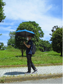 日差しが強い中、男性が小さめの青い日傘をさし、リュックを背負って歩道を歩いている写真