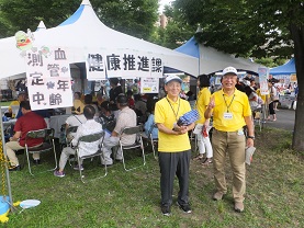 「健康推進課」「血管年齢測定中」と書かれたたくさんの来場者が訪れているテントの前に立つ2人の会員の男性の写真