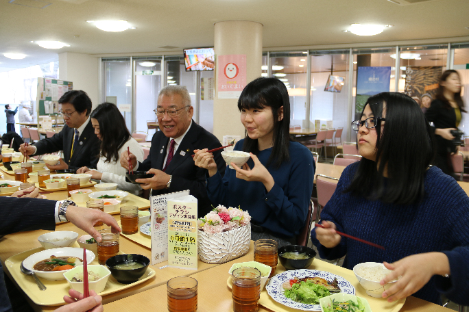 学校の食堂で、市長と学生の皆さんが同じテーブルに座り、食事を楽しんでいる様子の写真