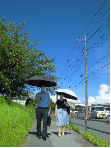 晴天の下、男性と女性が日傘をさしてカバンを持ち、歩道を歩いている写真
