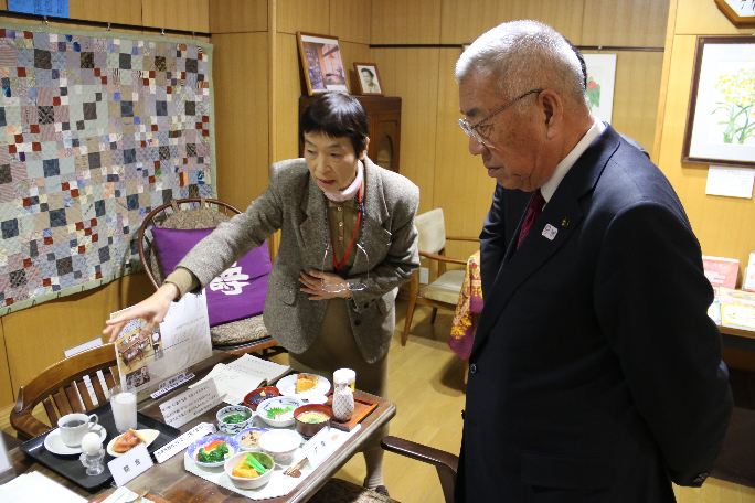テーブルに食品サンプルが並んでおり、職員の女性に説明を受けている市長の写真