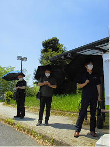晴天の下、3人の男性が青や黒の日傘をさしてカバンを持ち、バスを待っている写真