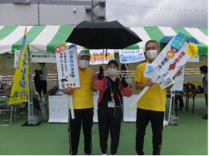中央に日傘をさした女性が立ち、その両側に黄色い服を着て熱中症予防啓発の立て札を持った男性が立ちピースをしている写真
