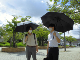 2人の男性が黒い日傘をさして樹木が植えられている遊歩道で立ち話している写真