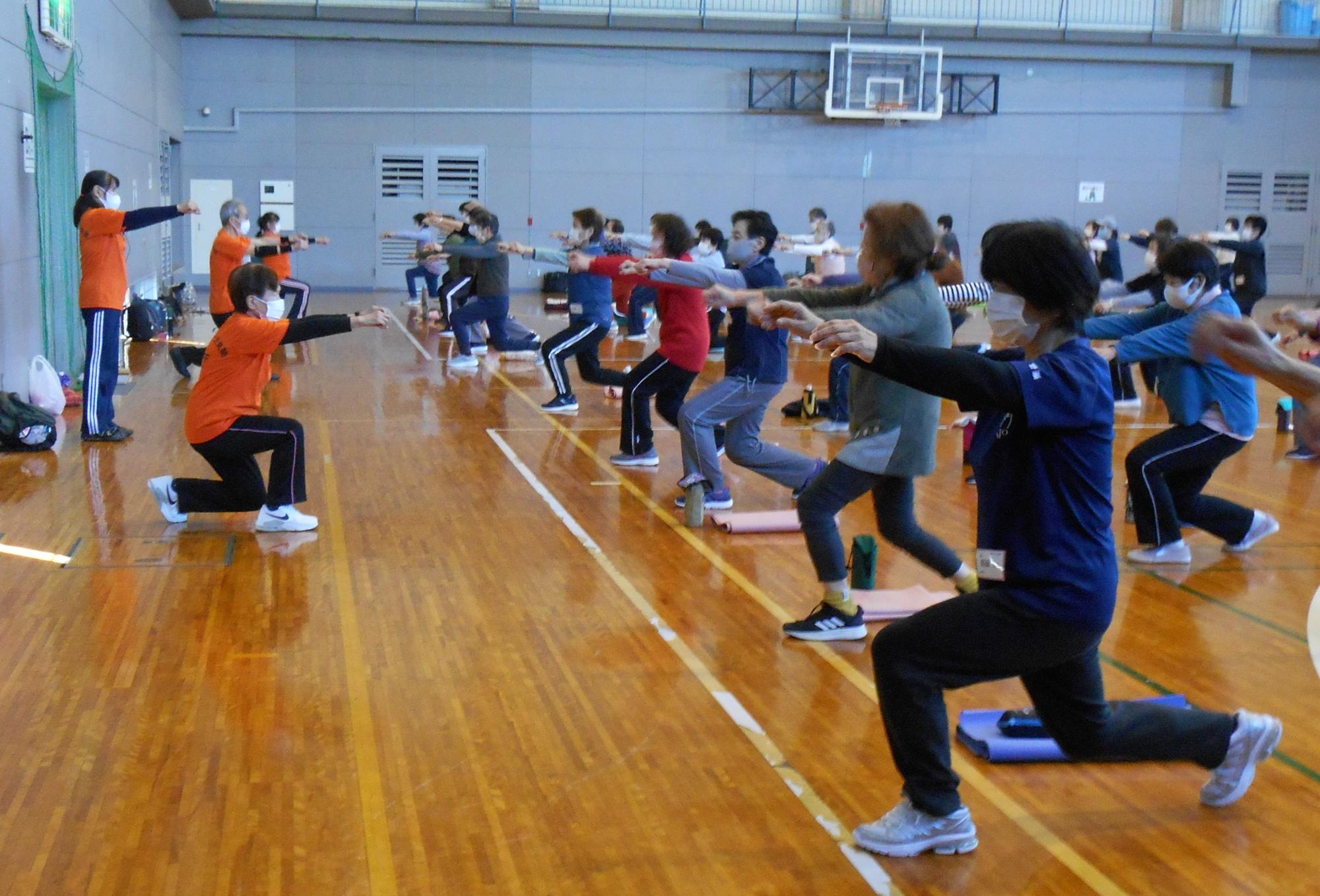 体育館で筋力トレーニング（ランジ）をしている4人のサポーターを見ながら、トレーニングをしている参加者の方々の写真