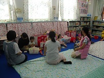 壁沿いに本棚が設置されている部屋でエプロン姿の関係者が見守る中、サロンに訪れた親子連れがおもちゃで遊んでいる様子の写真