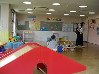 遊具がある室内の中央に赤ちゃんを寝かせ、女性1人が座り、1人が歩いているすまいるサロンの様子を写した写真