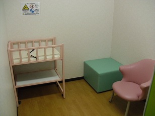 白い壁の部屋にベビーベッドと緑色の四角い椅子とピンクの椅子が置いてある写真