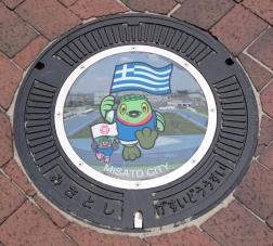 市のマスコットキャラクターかいちゃんとつぶちゃんがそれぞれギリシャの国旗と三郷市の市旗を持って走っているイラストが書かれたマンホール蓋の写真