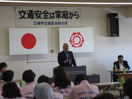 「交通安全は家庭から三郷市交通安全母の会」と書かれた横断幕と国旗と三郷市旗が飾られた室内に並んだ長机の席に座っている参加者と、向かい合って演壇に立っている市長が挨拶を行っている写真