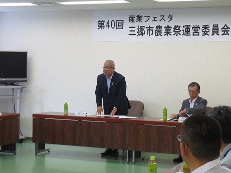 「第40回産業フェスタ三郷市農業祭運営委員会」と書かれた横幕が貼られた室内で、左側の席から立ち上がり挨拶を行っている市長の写真