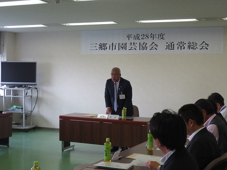 「平成28年度三郷市園芸協会通常総会」と書かれた張り紙が貼られた室内で、席から立ち上がり挨拶を行っている市長の写真