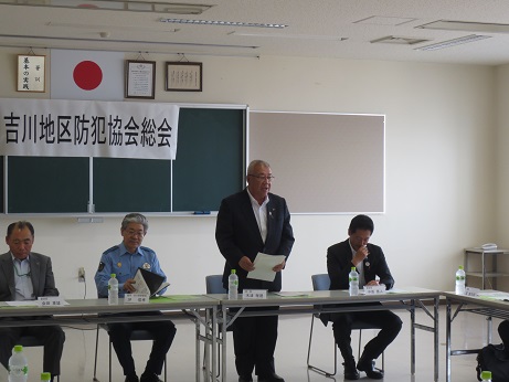 後方の黒板に「吉川地区防犯協会総会」と書かれた横幕が貼られた室内で、長机の席から立ち上がり挨拶を行っている市長の写真