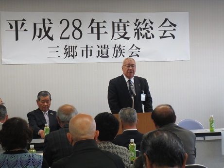 「平成28年度総会 三郷市遺族会」と書かれた横幕が壁に貼られた室内で、席から立ち挨拶を行っている市長の話を聞いている参加者の後ろ姿の写真