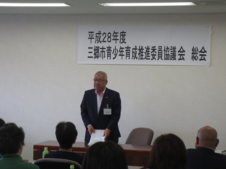 「平成28年度三郷市青少年育成推進委員協議会総会」と書かれた横幕が貼られた室内で、席から立ち上がり挨拶を行っている市長の写真