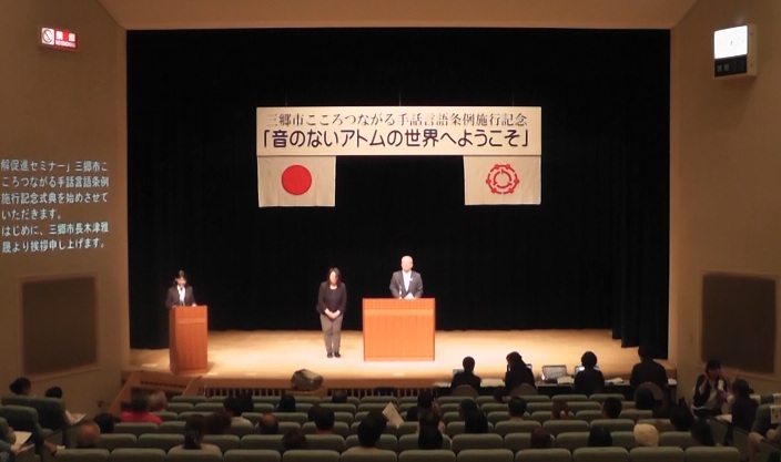 「音のないアトムの世界にようこそ」と書かれた横断幕と国旗、三郷市旗が吊り下げられているステージ上で、演台に立ってあいさつの言葉を述べる三郷市長と手話通訳の女性、観客席で話を聞く参加者の写真