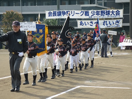野球場で旗を持った男性を先頭に続いてチーム名が記載されたプラカードを持ったユニホームを着た子供たちが一列に並んで入場行進を行っている写真