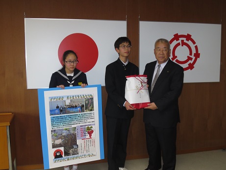 国旗と三郷市旗が飾られた室内で、左側の女子生徒が写真が入ったパネルを持ち、中央の男子生徒と市長が一緒に熨斗紙が貼られた赤色の包装紙で包まれた物を一緒に持っている写真